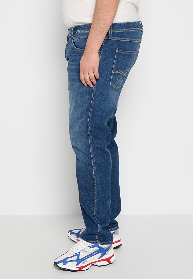 Blauwe denim jeans met een rechte pijp, vijf zakken en zichtbare stiksels. Draag je met witte sportschoenen met rode en blauwe accenten.