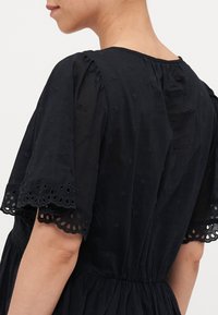 Svart bomullskjole med korte pufferm, scalloped blonderkant og broderte blomster mønstre. Rynket midje og teksturert finish.