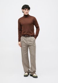 Junger Mann steht mit den Händen in den Taschen, trägt einen braunen Rollkragenpullover, beige Hosen und zweifarbige Lederschuhe vor einem schlichten weißen Hintergrund.