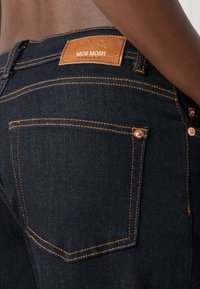 Primo piano di jeans in denim blu scuro con cuciture arancioni e una toppa in pelle marrone con l'etichetta MOS MOSH sulla fascia posteriore della vita.