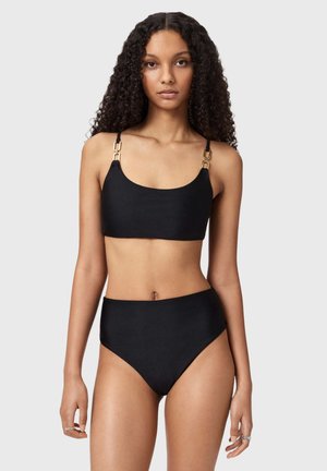 Kvinde med krøllet hår iført en sort bikini med guldspænder, stående mod en ensartet lys grå baggrund.