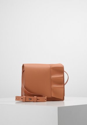 Petit sac bandoulière en cuir beige avec bandoulière ajustable, rabat pliant et détails froncés subtils sur un côté, présenté sur une surface blanche.