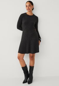 Frau, die ein dunkelgraues, geripptes Strickkleid und schwarze, spitze Stiefel bis zur Mitte der Wade trägt, steht vor einem schlichten, hellen Hintergrund.