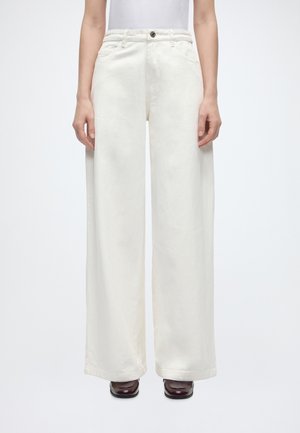 Personne portant un pantalon blanc taille haute à jambes larges avec fermeture par bouton et fermeture éclair, associé à des chaussures marron foncé à bout carré.