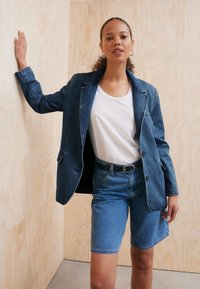 Blazer en denim avec des revers crantés, de couleur bleu foncé, et deux poches avant, porté sur un t-shirt blanc et un short en denim bleu clair.