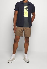Kortärmad marinblå skjorta med knappar över en grå t-shirt med en neongrön grafik, i kombination med beige shorts och vita sneakers.