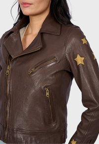 Veste de biker en cuir marron avec des accents en étoile dorée sur les manches. Comprend plusieurs fermetures éclair et un col cranté.