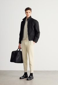 Czarna kurtka bomberka na jasnoszarym polo, kremowe spodnie o zwężanym kroju, czarne trampki i czarna torba sportowa w dłoni. Prosty, nowoczesny wygląd.