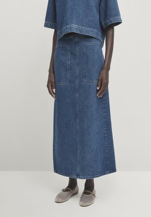 Modelka ubrana w ciemną spódnicę midi z denimu z łatami i kieszeniami, dopasowany krótki top z denimu oraz beżowe baleriny z paseczkami.