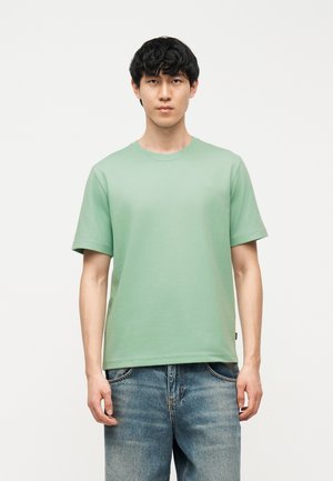 Giovane uomo in piedi, indossa una maglietta verde chiaro a maniche corte senza disegni e jeans blu, su sfondo bianco.