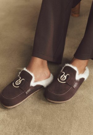 Person wearing dark brown fur-lined slip-on slippers with decorative metal buckle on beige carpet.