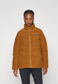 Giacca puffer in corduroy marrone con colletto alto e chiusura con zip. Presenta sezioni trapuntate orizzontali e un piccolo logo ricamato sul torace.