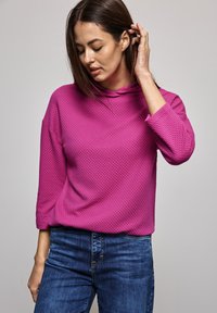 Sudadera texturizada en magenta con un corte relajado y cuello tipo embudo, combinada con jeans de mezclilla azules. La tela presenta un patrón de panal.