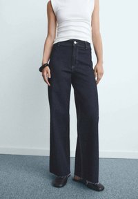 Donkerblauwe wijde jeans met een rafelige zoom, hoge taille en gele stikdetails. G gedragen met een witte, mouwloze top en zwarte schoenen.