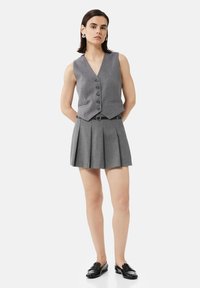 Gilet gris à boutons sur le devant, associé à une jupe plissée grise. Accentué par une ceinture noire. Le modèle porte des chaussures noires à enfiler.