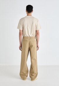 Beige Baumwoll-T-Shirt und locker sitzende khakifarbene Hose. Die Hose verfügt über zwei Gesäßtaschen und ein kleines Logo-Tag am Bund.