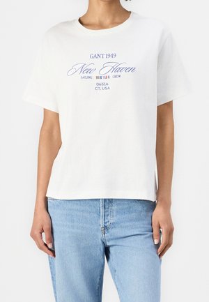 T-shirt blanc à manches courtes avec texte bleu "GANT 1949 New Haven Sailing Crew 06516 CT, USA" porté avec un jean bleu clair.