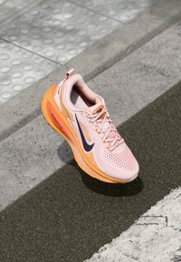 Zapatilla deportiva de color rosa claro con detalles en naranja, parte superior de malla, puntera redonda, logotipo negro, cordones blancos y suela acolchada, diseñada para correr.