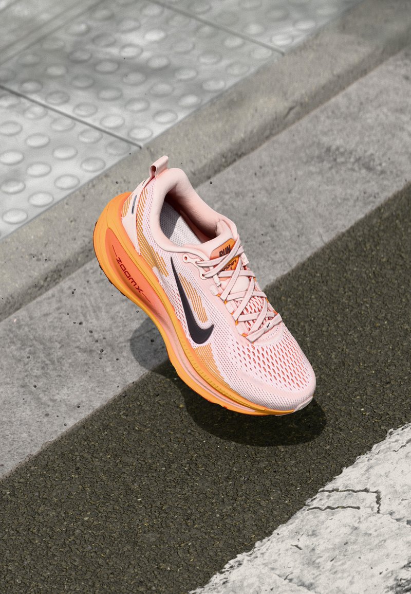 Zapatilla deportiva de color rosa claro con detalles en naranja, parte superior de malla, puntera redonda, logotipo negro, cordones blancos y suela acolchada, diseñada para correr.