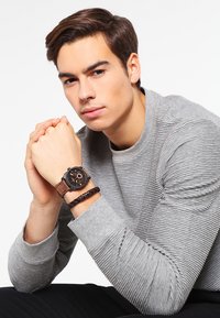 Bruine leren horloge met een zwarte wijzerplaat en zilveren accenten, gedragen om de pols; het model heeft ook een gevlochten zwarte armband. Grijze gemêleerde trui.