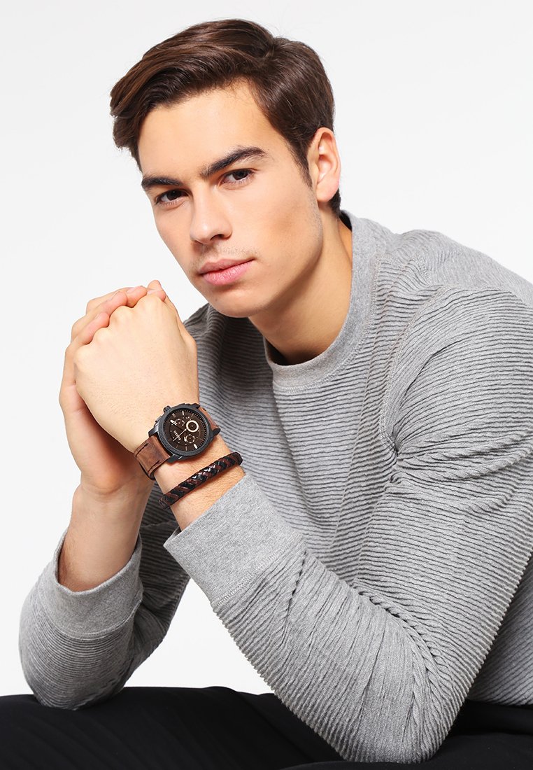 Bruine leren horloge met een zwarte wijzerplaat en zilveren accenten, gedragen om de pols; het model heeft ook een gevlochten zwarte armband. Grijze gemêleerde trui.