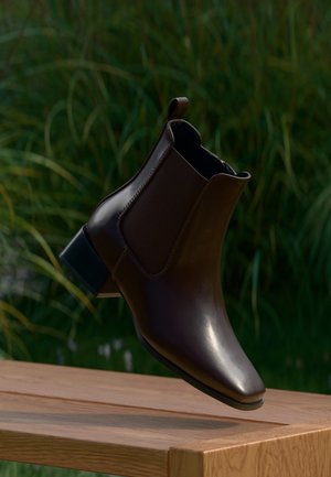 Stivaletto in pelle marrone scuro con tacco basso e linguetta, mostrato sospeso sopra una panca di legno con piante verdi sullo sfondo.