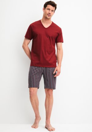 Mann steht barfuß, trägt ein rotes Kurzarm-V-Ausschnitt-Shirt und gestreifte Shorts vor weißem Hintergrund.