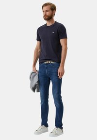 Maglietta a girocollo blu, jeans in denim blu, maglione grigio appoggiato sul braccio e sneakers bianche con accenti scuri. Design semplice e stile aderente.