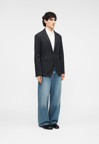 Blazer en chevron noir avec deux poches avant, porté sur une chemise blanche. Associé à un jean denim bleu clair ample et des chaussures noires.