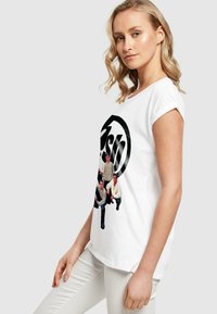 Tricou alb din bumbac cu un grafic circular negru și patru figuri îmbrăcate casual imprimate pe față. Mânecuțele sunt suflate.