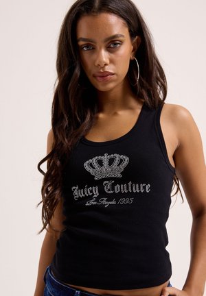 Frau mit langen dunklen Haaren trägt ein schwarzes Tanktop mit Strasskronen und der Aufschrift "Juicy Couture Los Angeles 1995", sichtbar sind auch Creolen.