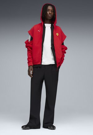 Junge Person mit mittellangen Dreadlocks trägt eine rote Puma-Ferrari-Jacke über einem weißen Hemd, schwarze Hosen und schwarze Schuhe.