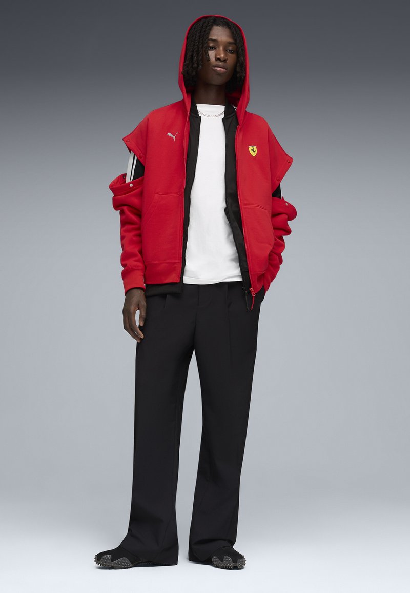 Unge med mellemlange dreadlocks bærer en rød Puma-Ferrari jakke over en hvid skjorte, sorte bukser og sorte sko.