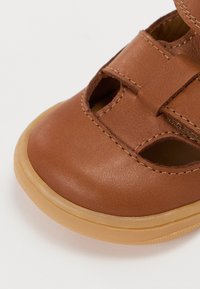 Hnědá kožená sandála s výřezovým designem, která má suchý zip a pružnou béžovou gumovou podrážku. Hladký povrch s prošívanými detaily.