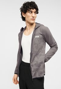 Γκρι φούτερ με φερμουάρ και καμουφλάζ σχέδιο, κατασκευασμένο από ελαφρύ υλικό. Διαθέτει κουκούλα και λογότυπο στο στήθος. Φοριέται πάνω από λευκό αμάνικο.