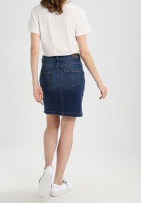 Džínová sukně v tmavě modré barvě, přiléhavý střih s kapsami na zadní straně, surový lem, kombinovaná s bílým tričkem a bílými teniskami, na světlém pozadí.