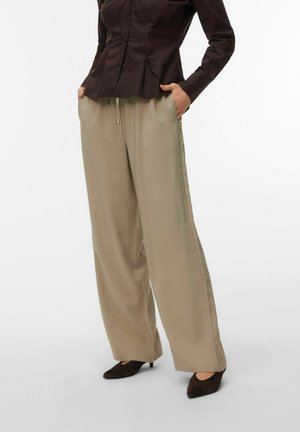 Donna che indossa pantaloni beige a gamba larga con coulisse in vita, camicia aderente marrone scuro con bottoni e tacchi scuri a punta, mani nelle tasche.