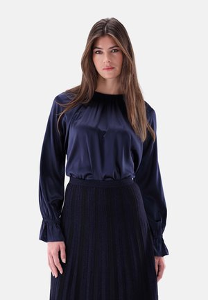 Navy Satinbluse mit glatter Textur, gerafftem Ausschnitt und langen Puffärmeln. Kombiniert mit einem plissierten dunklen Rock, der einen dezenten Glanz aufweist.