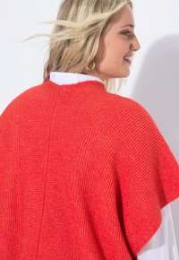 Röd stickad poncho med ett texturerat stickmönster, med rund halsringning och lös passform, lager över en vit skjorta.