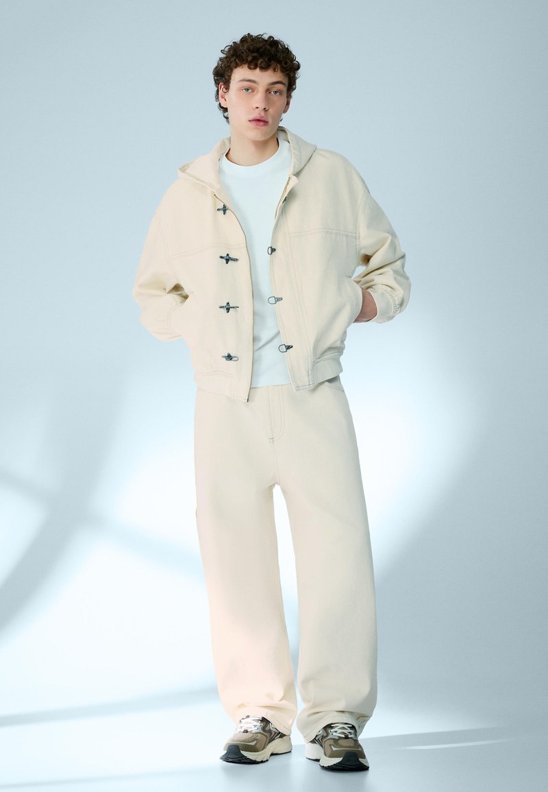 Ung person med lockigt hår som bär en beige huva, vit skjorta, vida beige byxor och gråa sneakers, står mot en ljusblå bakgrund.