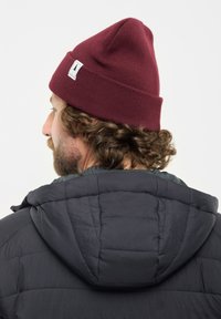 Uomo con capelli ricci e barba che indossa un berretto bordeaux e un giubbotto nero trapuntato con cappuccio, rivolto a sinistra su uno sfondo semplice.