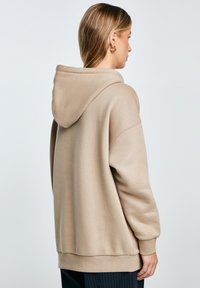Béžová mikina s kapucí s volným střihem, měkkou texturou a žebrovanými manžetami. Hladká látka s minimálním zdobením, zobrazeno ze zadní strany.