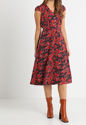 Vrouw draagt een zwarte midi-jurk met rood bloemenpatroon en kapmouwtjes, gecombineerd met glanzende bruine enkellaarsjes met hak.