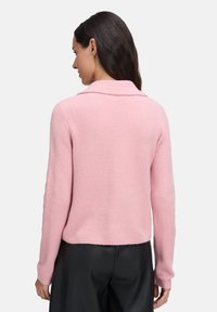 Frau mit langen dunklen Haaren, die einen weichen, pinken Langarmpullover und schwarze Hosen trägt, von hinten vor einem schlichten weißen Hintergrund. 