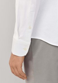 Lewe ręka obok szarych spodni, na sobie biała koszula z długim rękawem z zapięciem na guziki i teksturowanym detalem materiału.