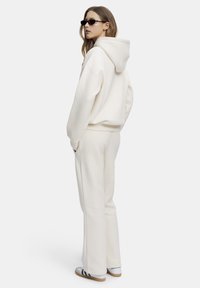 Krämfärgad huvtröja med en stor huva, kombinerad med matchande krämfärgade byxor. Accentuerad med svarta solglasögon och vita sneakers med gröna ränder.