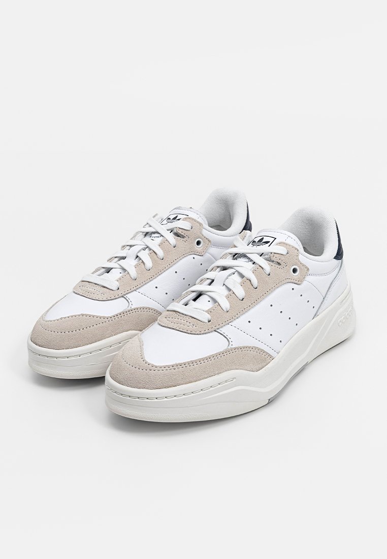Zapatillas Adidas blancas y beige con cordones, paneles laterales perforados y detalles en negro en el talón, mostradas una al lado de la otra sobre un fondo blanco.