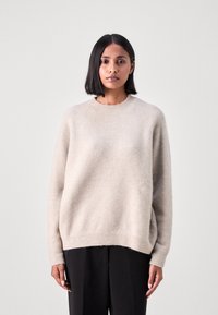 Maglione lavorato a maglia color beige chiaro, realizzato in materiale morbido, con scollatura rotonda, spalle scese e vestibilità comfortevole. Abbinato a pantaloni scuri.