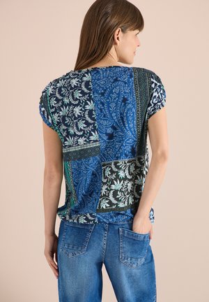 Femme de dos portant un haut à manches courtes composé d'un patchwork à motifs floraux et cachemire bleus, et un jean en denim bleu, une main dans la poche arrière.