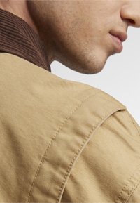 Beez kollane jakk, mille õlgadel on nähtav tekstuur ja õmblused; pruun kašmiirkaelus lisab kontrastset elementi.